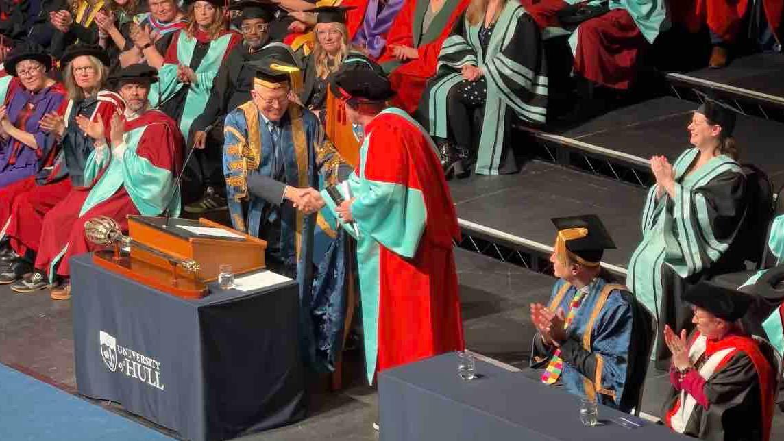 An audience applauding Radford while he is shaking the Dean's hand on the stage.  