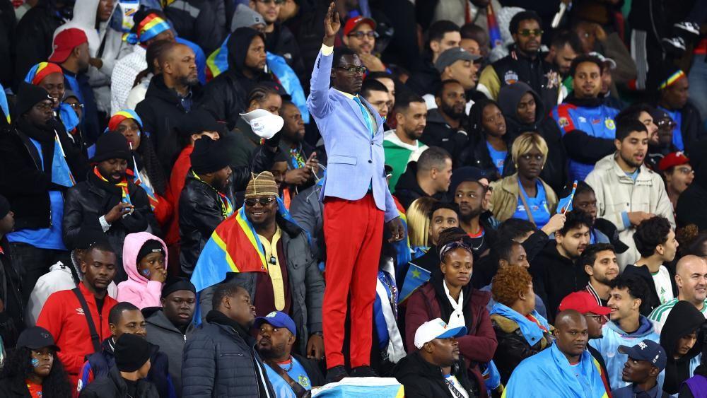 Michel Nkuka Mboladinga dressed as Patrice Lumumba in a blue suit jacket and red trousers with his hand raised in the middle of Congolese fans in a stadium in Rabat.
