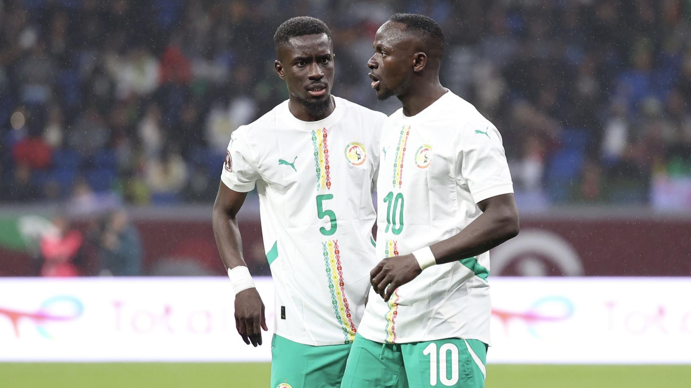 Idrissa Gana Gueye stands talking to Sadio Mane during a football match. Both players wear white shirts bearing green, yellow and red detail on the front and green shorts. Gana Gueye's shirt has the the number five on the chest while Mane's kit bears a 10 on the chest and left short leg
