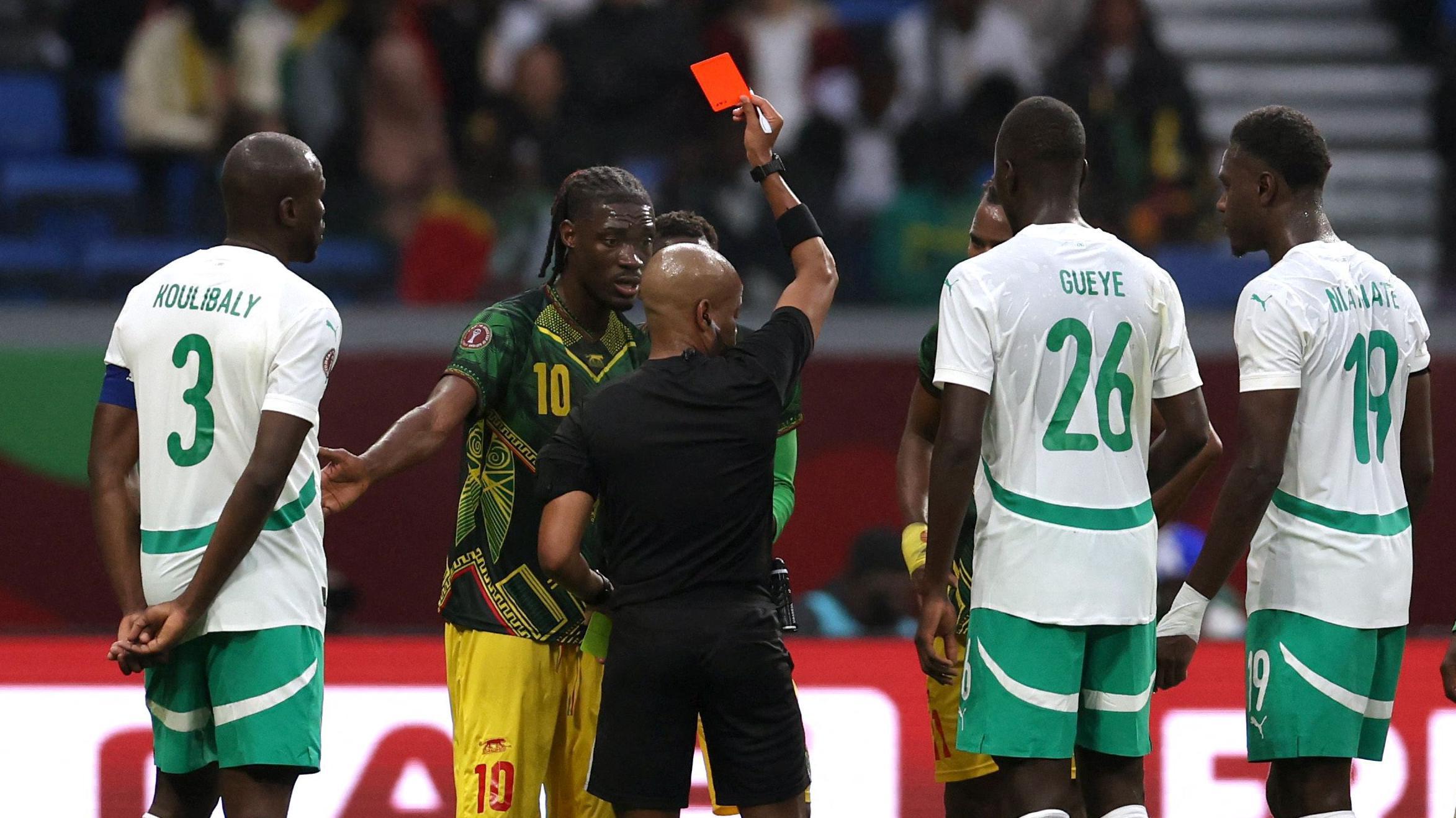 Referee Abongile Tom, dressed all in black, is seen from behind raising a red card aloft towards Mali's Yves Bissouma, who is wearing a green shirt and yellow shorts. Three Senegal players in white shirts and green shorts are seen from behind looking on. Blurred in the background is a stand with blue seats partially occupied by fans