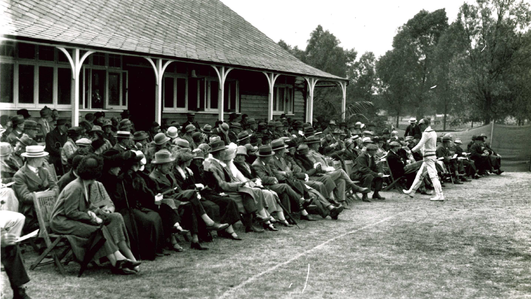 A black and white image from Essex against Oxford University match. Several rows of people form the crowd in front of a clubhouse. They are dressed smartly with hats on. A cricketer wearing whites walks past them.